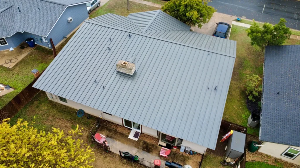 Angled drone view of a gray standing seam metal roof with a stone chimney and cricket on a single-story home, beside a neighbor's shingle roof