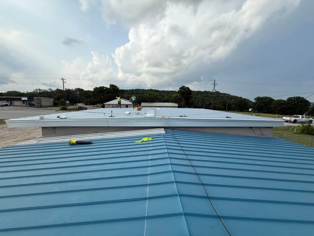 Roofing crew installing new silver metal panels on the upper flat section of a commercial building in Burnet, TX