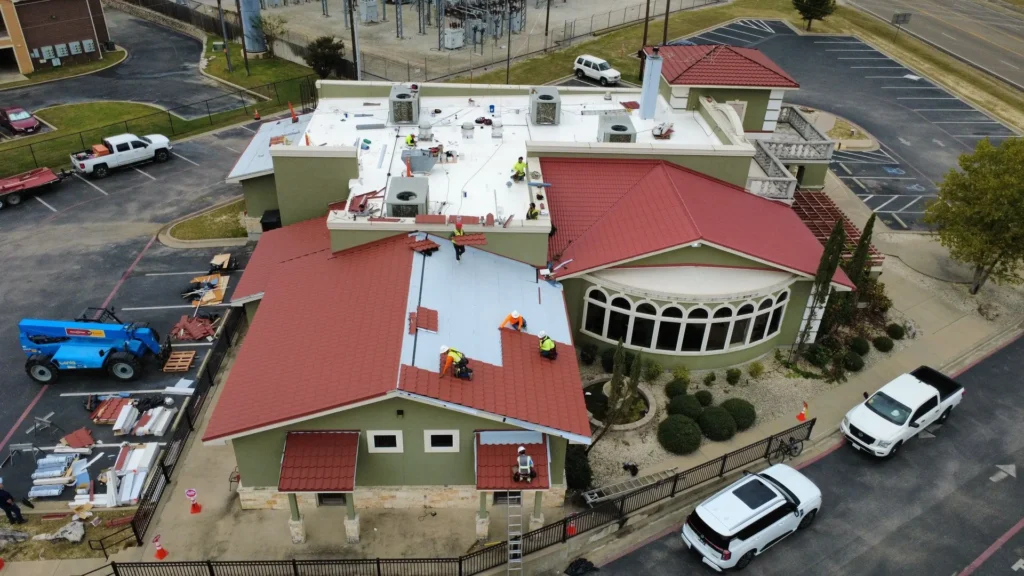 Roofing crews installing metal tile panels over new underlayment while another crew works on the PVC flat roof, with a staged material area and telehandler in the parking lot