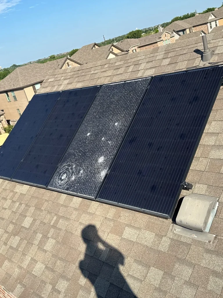 A shattered rooftop solar panel with visible hail impact marks beside undamaged panels on an asphalt shingle roof.