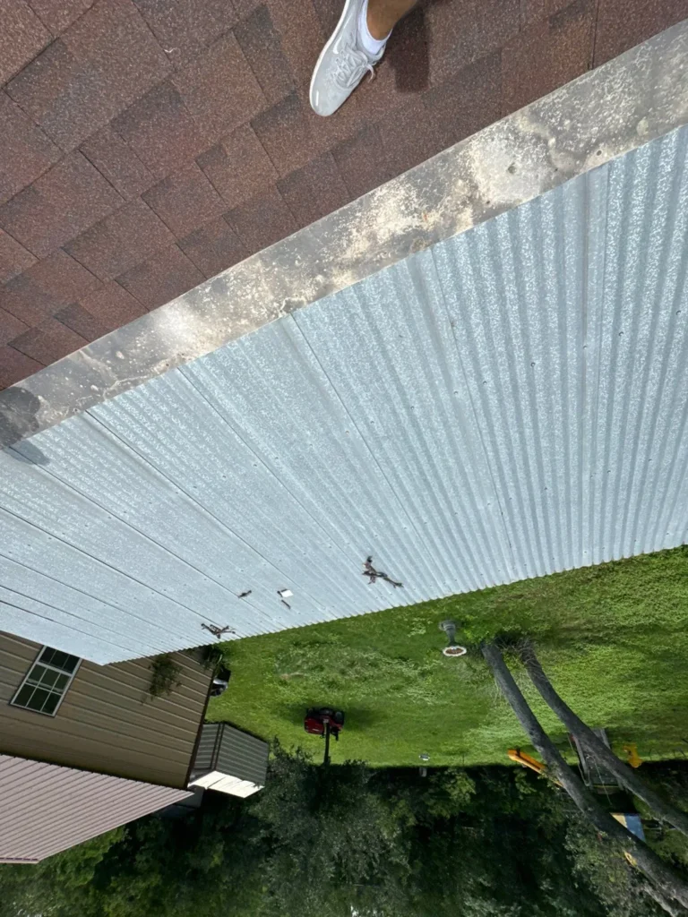 Old galvanized corrugated metal patio roof with debris and worn flashing next to the shingle roof in Helotes