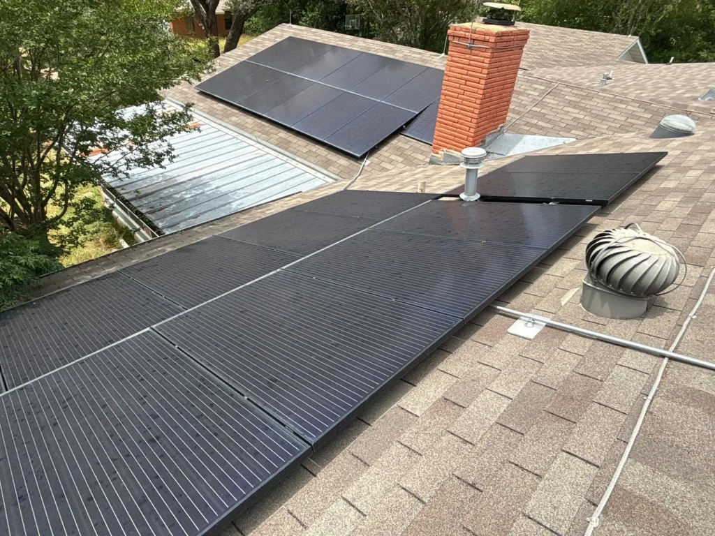 Multiple black solar panels mounted across an asphalt shingle roof with vents, flashing and a brick chimney visible.