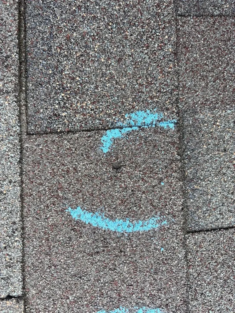 Close-up of a hail impact on an asphalt shingle with the mat exposed, circled in blue chalk in Uhland, TX